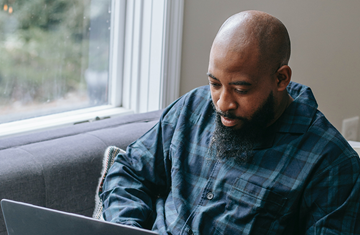 Black man sitting on sofa next to a window working on his laptop