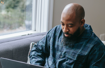 Black man sitting on sofa next to a window working on his laptop